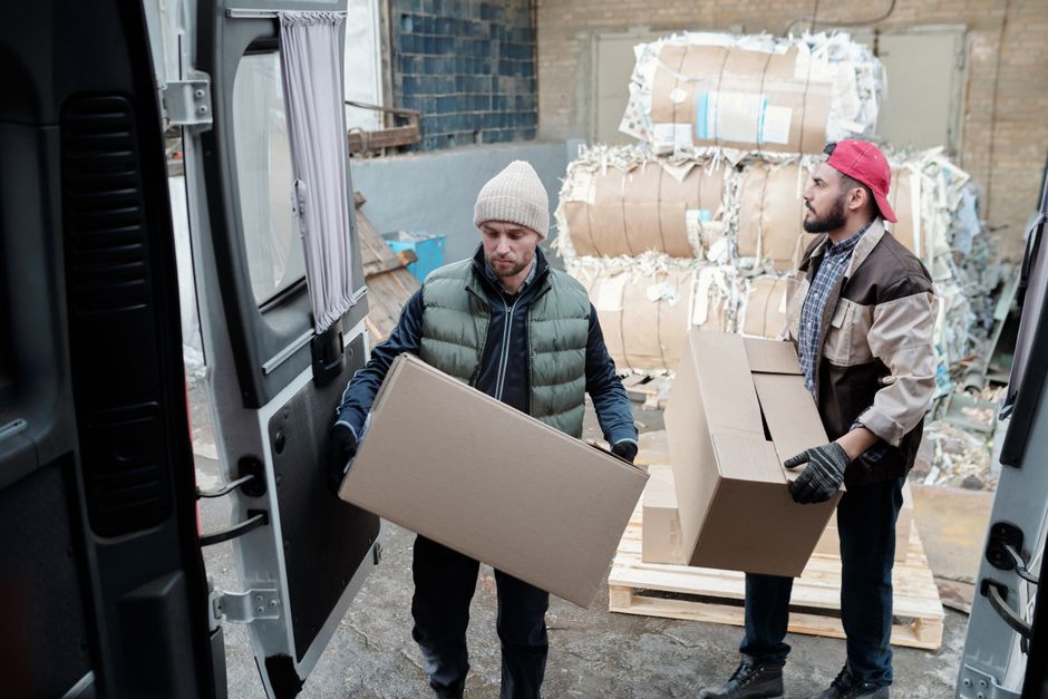 Two workers loading flattened cardboard boxes into a van at a recycling yard, with baled waste paper stacked behind them
