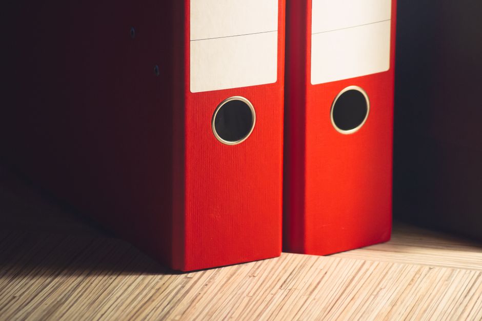 Red lever-arch files with blank spine labels standing on a shelf, typical of office archive storage