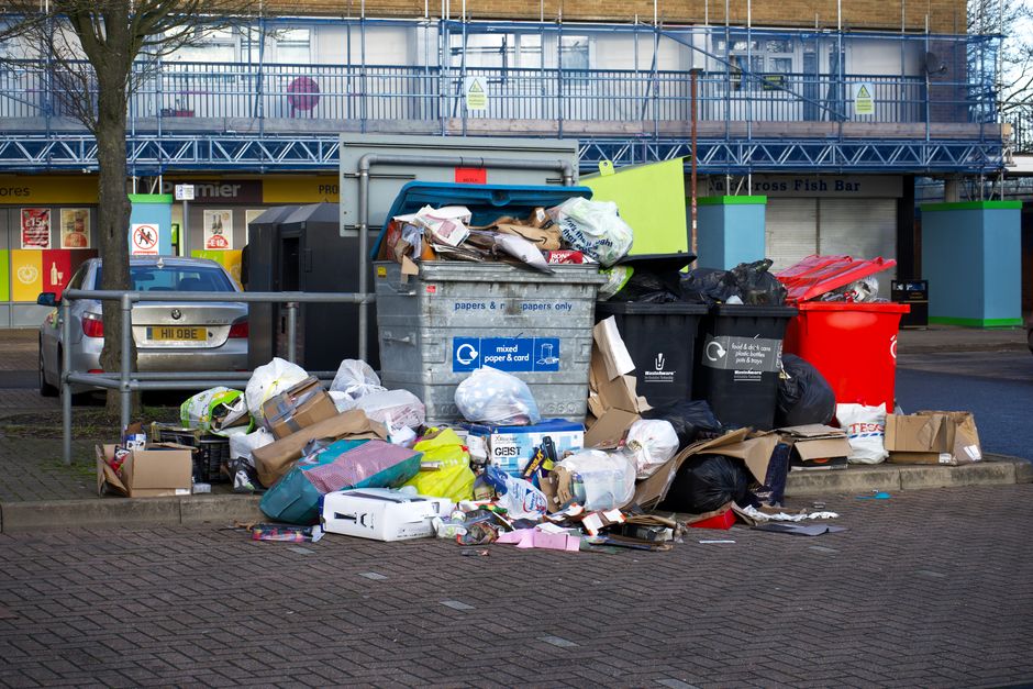 Overflowing bins and bagged rubbish piled up on a paved area outside shops on a UK street