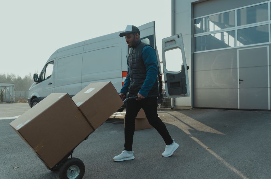 Man pulling a sack truck loaded with cardboard boxes towards an open van outside a warehouse