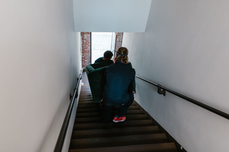 Two people carrying a sofa down an indoor staircase during a collection