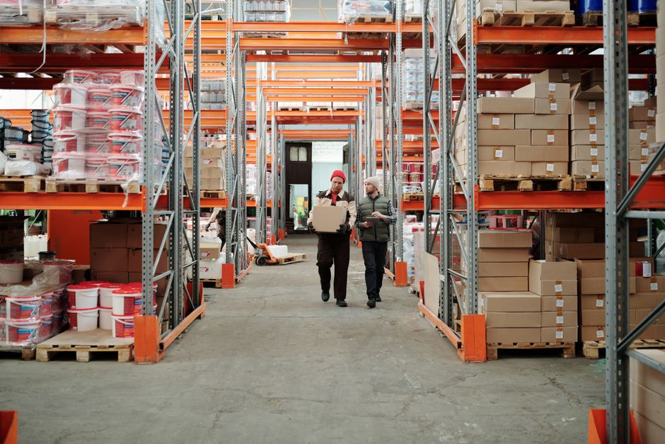 Two warehouse workers carrying a box down an aisle between loaded pallet racking during a stock clearance